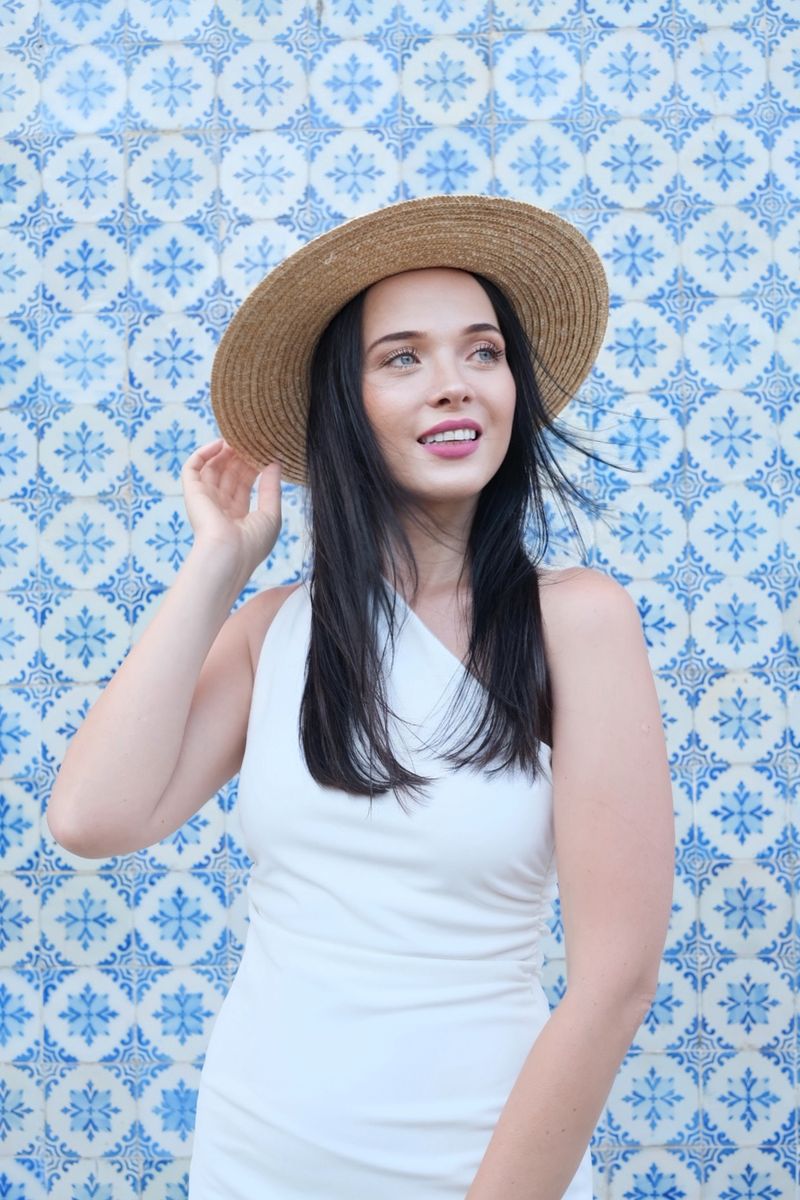 Karolina Jakubowicz in a white dress and straw hat in front of a blue and white azulejo tiled wall